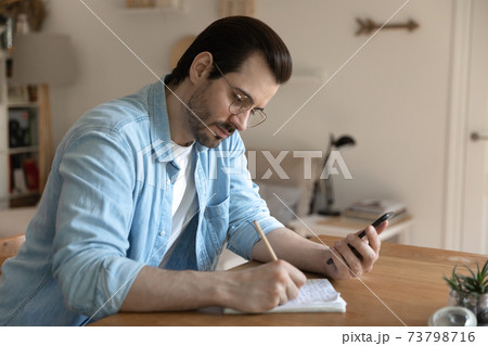 Young male student watching distant lecture by smartphone making notes Young male student watching distant lecture by smartphone making notes 73798716
