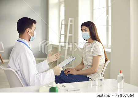 Doctor and patient in medical face masks sitting at table at the hospital and talking 73807064