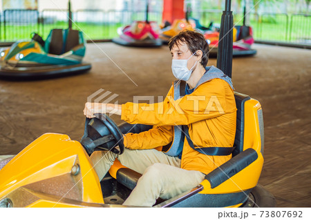Man wearing a medical mask during COVID-19 coronavirus having a ride in the bumper car at the amusement park 73807692