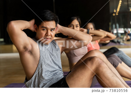 Group of Asian women and man doing abdominal exercises on yoga mats in aerobics class. Young sporty people twisting body on the floor in gym studio. Group of Asian women and man doing abdominal exercises on yoga mats in aerobics class. Young sporty people twisting body on the floor in gym studio. 73810556