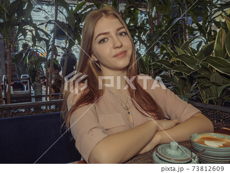 Blond girl in cafeteria near the potted plants with coffee in front of her looking at camera. Cafe city lifestyle woman drinking coffee. 73812609