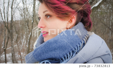 Attractive young woman with perfect skin and makeup with dark pink hair is possing on winter park background wearing blue scarf and silver earrings 73832813