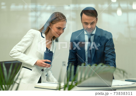 Smiling millennials diverse taking coffee break looking at laptop monitor sitting behind a glass wall in the modern office. Partners discussing project sitting in a modern bright meeting room Smiling millennials diverse taking coffee break looking at laptop monitor sitting behind a glass wall in the modern office. Partners discussing project sitting in a modern bright meeting room 73838311