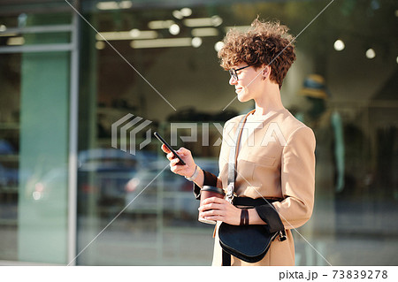 Curly brunette businesswoman with drink and handbag messaging in smartphone 73839278