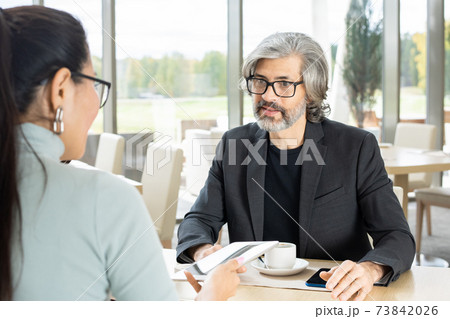 Serious mature businessman answering question of his young female colleague Serious mature businessman answering question of his young female colleague 73842026