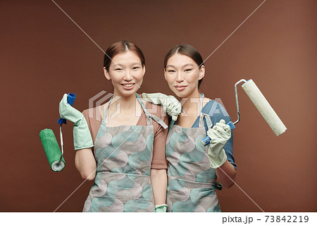 Happy young Asian female twins in aprons and gloves holding paint rollers 73842219