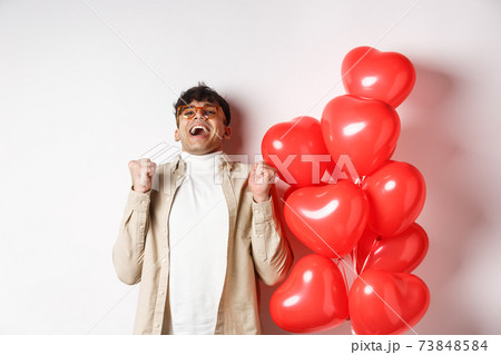 Valentines day. Happy modern man celebrating, screaming from joy and happiness, having date with lover, being in love, standing near heart balloons on white background 73848584