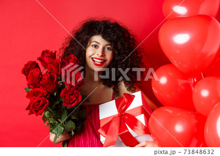 Happy Valentines day. Romantic girl with presents from lover, holding bouquet of roses and gift box, standing near cute red hearts on studio background 73848632