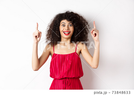 Happy elegant woman in red dress and makeup, smiling amused and pointing fingers up at logo, showing advertisement, standing over white background Happy elegant woman in red dress and makeup, smiling amused and pointing fingers up at logo, showing advertisement, standing over white background 73848633