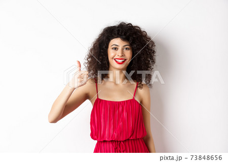 Cheerful woman with red lips and elegant dress, showing thumb up and smiling, recommending product, standing over white background 73848656