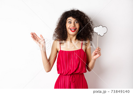 Happy young woman in red dress, holding comment cloud and looking surprised, standing over white background Happy young woman in red dress, holding comment cloud and looking surprised, standing over white background 73849140