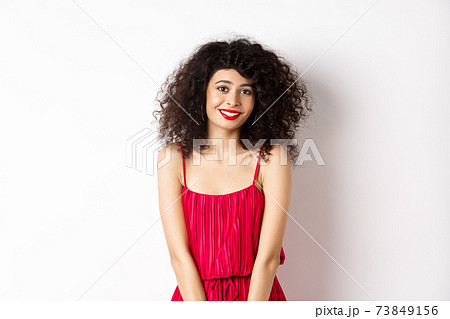 Cute modest lady in red dress and makeup, smiling and blushing, looking at camera, standing over white background Cute modest lady in red dress and makeup, smiling and blushing, looking at camera, standing over white background 73849156