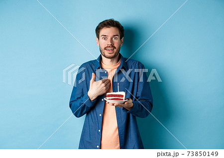Celebration and holiday concept. Surprised guy holding birthday cake with candle and pointing at himself with disbelief, being congratulated with b-day, blue background 73850149