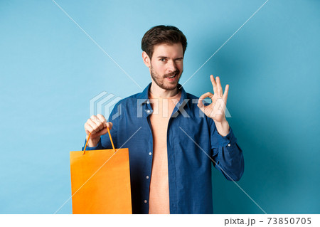 Modern man in casual outfit showing shopping bag and okay sign, winking at camera, recommending shop, standing on blue background 73850705