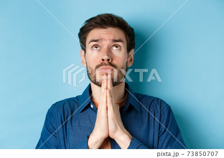 Close up shot of hopeful guy pleading God, holding hands in begging gesture and looking up in sky, standing on blue background supplicating 73850707