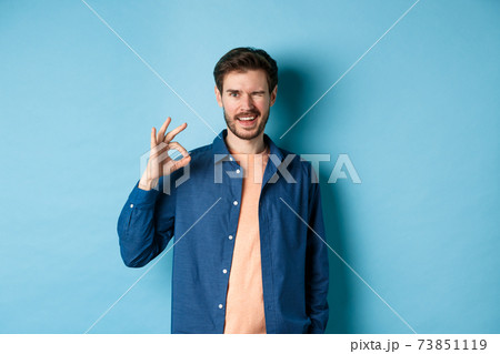 Positive smiling guy winking and showing okay sign, praising good work, recommending good company, standing on blue background Positive smiling guy winking and showing okay sign, praising good work, recommending good company, standing on blue background 73851119
