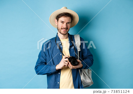 Handsome guy going on summer vacation, backpacking on holiday. Tourist with straw hat and camera smiling happy, standing on blue background Handsome guy going on summer vacation, backpacking on holiday. Tourist with straw hat and camera smiling happy, standing on blue background 73851124