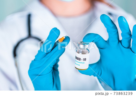 Woman doctor or nurse in uniform and gloves wearing face mask protective in lab holding medicine vial vaccine bottle with COVID-19 Coronovirus vaccine label 73863239