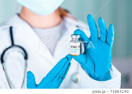Woman doctor or nurse in uniform and gloves wearing face mask protective in lab holding medicine vial vaccine bottle with COVID-19 Coronovirus vaccine label 73863240