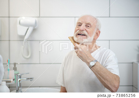 Elderly man in white tshirt brushing his beard Elderly man in white tshirt brushing his beard 73865256