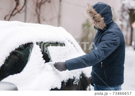 Young man cleaning snow from car windshield outdoors near the garage on winter day Young man cleaning snow from car windshield outdoors near the garage on winter day 73866657