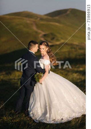 Full length body portrait of attractive bride and stylish groom enjoying romantic moments in the mountains at sunset in beautiful summer day. Wedding couple. Green hills on background 73866915