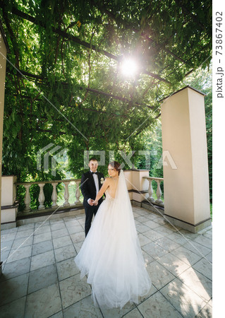 Wedding couple of groom and bride stand on big gorgeous balcony. Sun rays through the leafs 73867402
