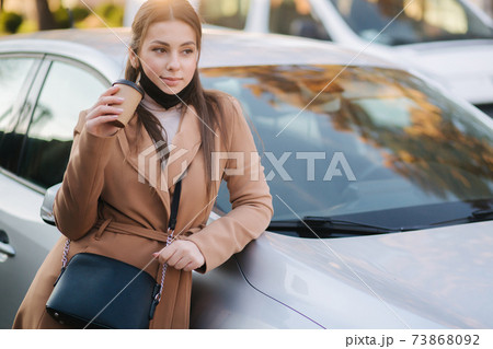Beautiful young woman in black face mask stand by the car and drink coffee. Happy young woman have a break. Female pulled off mask for drinking cappucino outdoors Beautiful young woman in black face mask stand by the car and drink coffee. Happy young woman have a break. Female pulled off mask for drinking cappucino outdoors 73868092