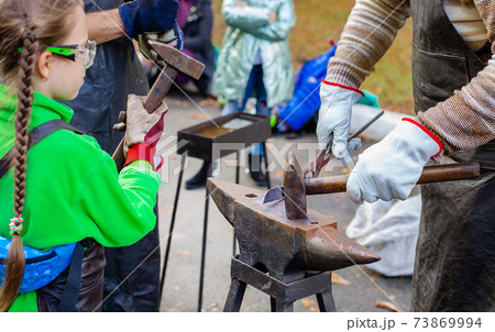 A blacksmith forges a metal blank on the anvil at a fair in the presence of spectators. A blacksmith forges a metal blank on the anvil at a fair in the presence of spectators. 73869994