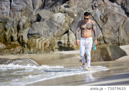 Athletic tattooed guy walking at the tropical beach Athletic tattooed guy walking at the tropical beach 73879620