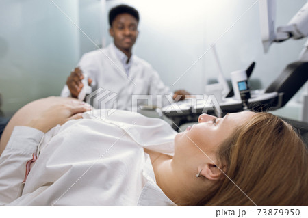 Cropped close up of young pregnant woman, visiting her male African doctor sonographer, having ultrasound scanning at the medical clinic. Ultrasound equipment, technology concept Cropped close up of young pregnant woman, visiting her male African doctor sonographer, having ultrasound scanning at the medical clinic. Ultrasound equipment, technology concept 73879950