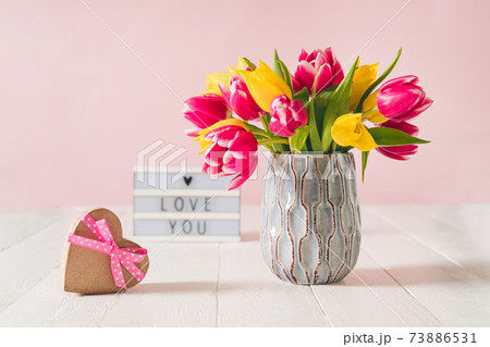 Recycled Heart shape gift box, spring bouquet of pink and yellow tulip flowers in a vase and Lightbox with love you message on the white wooden table and pink background. Greeting card mockup. Recycled Heart shape gift box, spring bouquet of pink and yellow tulip flowers in a vase and Lightbox with love you message on the white wooden table and pink background. Greeting card mockup. 73886531