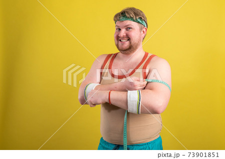 A funny, cheerful fat man in sportswear measures his arms. Overweight man goes in for sports. Yellow background isolate 73901851