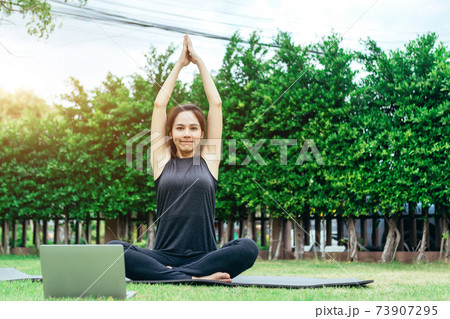 Relaxed healthy sports Asian woman in fitness clothes at park meditating using online streaming yoga site in laptop. 73907295