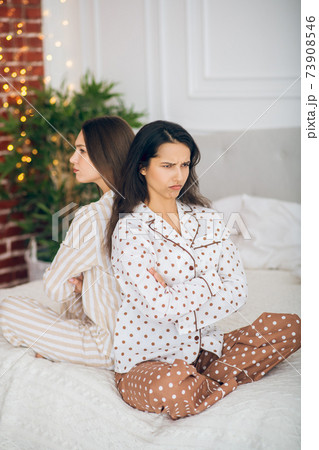 Two young girls in pajamas sitting back to back on a bed and looking moody 73908546