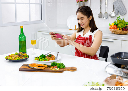 Asian woman in red apron taking a picture of food she make with mobile phone. Concept woman preparing meals at home. Asian woman in red apron taking a picture of food she make with mobile phone. Concept woman preparing meals at home. 73916390