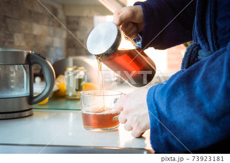 Close up man in home bathrobe pouring hot tea from glass teapot into cup on the kitchen table. Process brewing tea in the morning. A cup of freshly brewed black tea, warm soft light. Selective focus. Close up man in home bathrobe pouring hot tea from glass teapot into cup on the kitchen table. Process brewing tea in the morning. A cup of freshly brewed black tea, warm soft light. Selective focus. 73923181