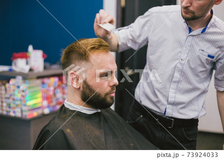 Barber Making Haircut Bearded Man In Barbershop. Professional stylist cutting client hair in salon. Barber using scissors and comb. Skillful hairdresser cutting male hair. Hair Care Service Concept Barber Making Haircut Bearded Man In Barbershop. Professional stylist cutting client hair in salon. Barber using scissors and comb. Skillful hairdresser cutting male hair. Hair Care Service Concept 73924033