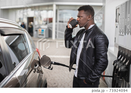 Side view of young African man, standing outdoors at the gas station, drinking take away coffee while refueling his luxury car's tank by petrol pump nozzle Side view of young African man, standing outdoors at the gas station, drinking take away coffee while refueling his luxury car's tank by petrol pump nozzle 73924094