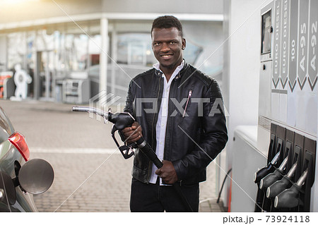 Young African man wearing casual outfit, is ready to refueling his luxury car, holding filling gun nozzle at the gas station, looking at the camera with smile 73924118