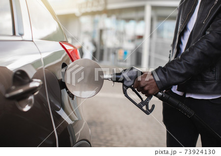 Cropped close up image of unrecognizable African businessman in casual wear, rholding in hand filling gun and refueling his luxury car with gasoline at the petrol station Cropped close up image of unrecognizable African businessman in casual wear, rholding in hand filling gun and refueling his luxury car with gasoline at the petrol station 73924130