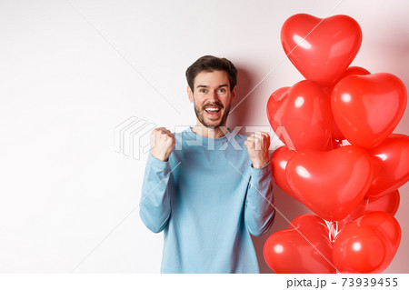 Portrait of happy guy celebratiing lovers day, standing near valentines red heart balloon and cheering, making yes gesture and smiling at camera, standing over white background 73939455