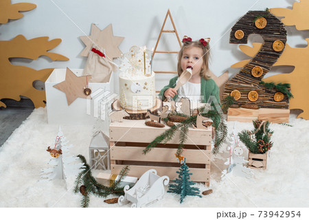Children's winter birthday decorations. 2 years. Cute little girl in a green cardigan near white birthday cake Children's winter birthday decorations. 2 years. Cute little girl in a green cardigan near white birthday cake 73942954