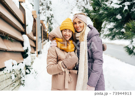 Two happy laughing young friendly women stand on a snowy street in a European rural suburb. Two happy laughing young friendly women stand on a snowy street in a European rural suburb. 73944913