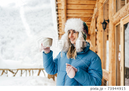 woman in a hat with earflaps with a mug of drink on the background of the chalet woman in a hat with earflaps with a mug of drink on the background of the chalet 73944915