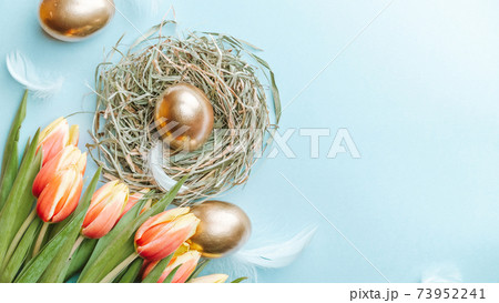 Easter eggs Golden color in basket with spring tulips, white feathers on pastel blue background in Happy Easter decoration. Festive decoration in flat lay. 73952241