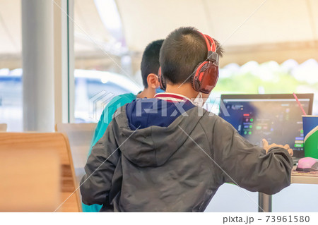 Back view of Asian boy wearing protective face mask and headphones while working with a friend on laptop during corona virus (Covid-19) and flu outbreak in food court at the mall.New normal lifestyle. 73961580