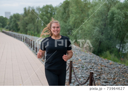 An obese young woman jogging outdoors. Fat beautiful smiling girl in a black tracksuit is engaged in fitness for weight loss on the waterfront. A woman runs on a summer day. An obese young woman jogging outdoors. Fat beautiful smiling girl in a black tracksuit is engaged in fitness for weight loss on the waterfront. A woman runs on a summer day. 73964627