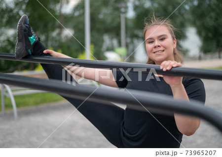 Fat girl is engaged in stretching on the uneven bars on the playground. Beautiful young smiling overweight woman doing sports for weight loss outdoors. 73965207