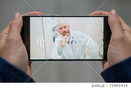 Closeup of female hands with a phone. A bald man doctor remotely consults a patient with a webcam. The woman says with a friendly physician for video calls. 73965414